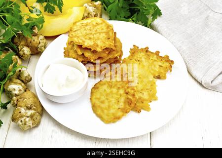 Pumpkin and Jerusalem artichoke fritters with sour cream in a plate, napkin, parsley on light wooden board background Stock Photo