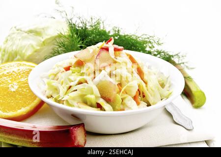 Fresh cabbage, carrot and rhubarb salad with orange juice, honey and mayonnaise dressing in a plate on a napkin, dill and fork on light wooden board b Stock Photo