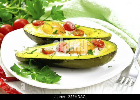 Scrambled eggs with cherry tomatoes in two halves of avocado in a plate, green towel and fork on the background of white wooden board Stock Photo