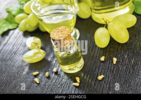 Grape oil in a bottle, a gravy boat and a jar, berries and seeds of green grapes on the background of dark wooden board Stock Photo