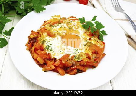 Fried eggs with tomatoes, sweet pepper, onions and herbs in a plate, napkin, parsley and fork on background of light wooden board Stock Photo