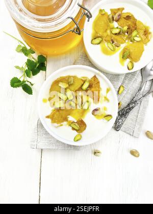 Dessert of yogurt, persimmon and honey with vanilla, cardamom and pistachios in two bowls on a napkin, mint and spoon on wooden board background from Stock Photo