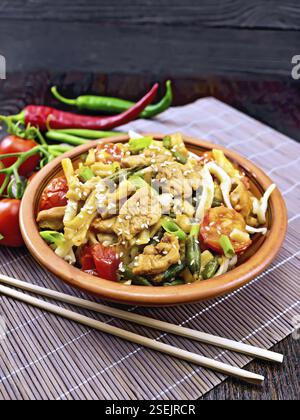 Thai noodles wok with chicken meat, tomatoes, soy sauce and green beans sprinkled with sesame seeds in a plate on a bamboo napkin with chopsticks on a Stock Photo