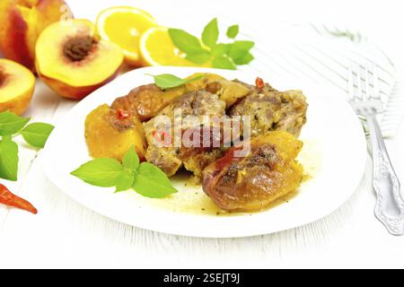 Turkey stewed with peaches, fresh hot pepper and orange sauce, basil leaves in a plate, dishcloth, fruits on wooden board background Stock Photo