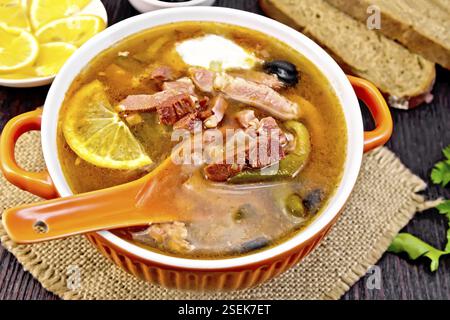 Soup saltwort with lemon, meat, pickles, tomato sauce olives, spoon in a bowl on a napkin of burlap on the background of a wooden board Stock Photo