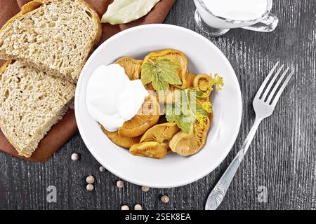 Salted mushrooms saffron with sour cream, currant leaf and sprig of dill in plate, fork, napkin and bread on wooden board background from above Stock Photo