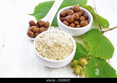 Flour and hazelnuts in two bowls, a spoon with peeled walnut kernels and a branch of filbert with leaves on the background of light wooden board Stock Photo