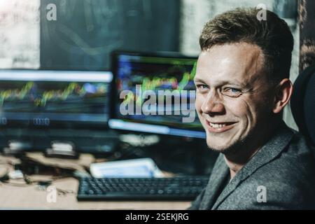Man smiling in front of computer screens displaying colorful trading charts Stock Photo