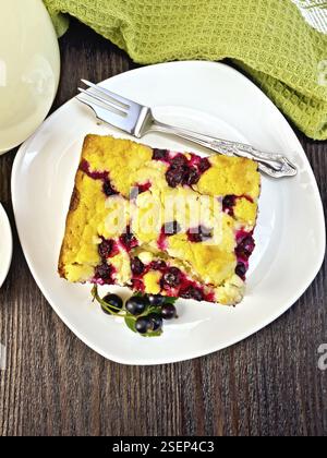 Piece of sweet pie with black currant berries and fork on a plate, napkin, milk in a glass jug on a wooden board background from above Stock Photo