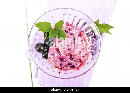 Ice cream with black currant in a glass goblet on a lilac napkin, berries with leaves on a wooden plank background from above Stock Photo