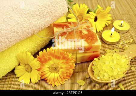 Two bars of homemade soap, towels, bath salts in a wooden spoon, two candles and marigold flowers on a background of bamboo napkins Stock Photo
