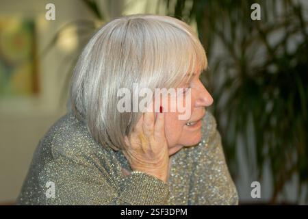 An elderly woman with silver hair and a thoughtful expression rests her head on her hand while engaged in conversation. Stock Photo