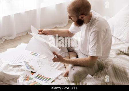 Accounting bookkeeping concept. Busy bearded man works at home on bed with paper documents in bedroom interior. Stock Photo