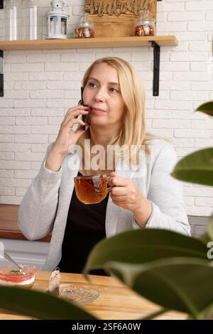 A stylish young woman sits at a cafe table, talking on her phone while enjoying a cup of tea. She smiles and looks off into the distance, surrounded b Stock Photo