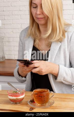 Elegant middle-aged woman sitting at the kitchen table, looking at her smartphone with a thoughtful expression. In front of her is a cup of tea and a Stock Photo