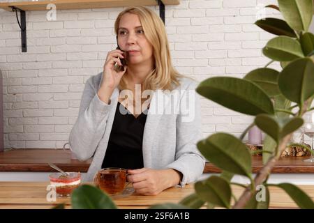 Stylish middle-aged woman in gray suit sitting at cafe table, talking on phone, holding a cup of tea, looking thoughtful. Dessert and plants visible i Stock Photo