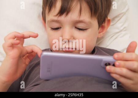 A close-up of a child using a mobile phone while lying in bed. A toddler exploring a mobile phone while resting. Kid aged three (three-year-old ) Stock Photo