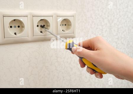Hand using screwdriver to repair electrical socket on patterned wallpaper background. Home maintenance and electrical work concept. Stock Photo
