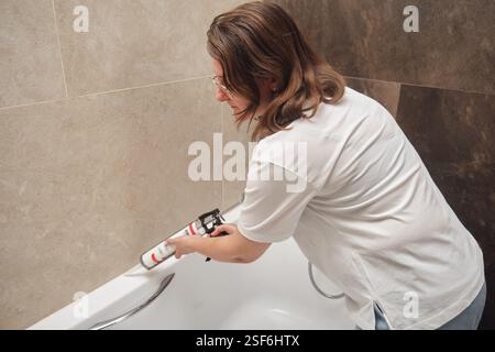 Woman applying sealant to bathtub corner using caulking gun. Home renovation and bathroom maintenance concept. Stock Photo