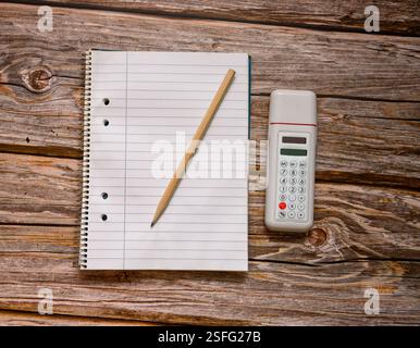 A spiral notebook lies open beside a corded calculator on a rustic wooden table. A yellow pencil rests next to the notebook Stock Photo