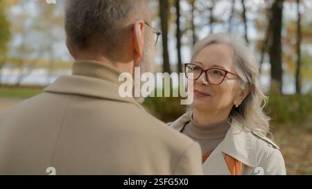 Gray-haired 60s mature Caucasian woman wife embrace cuddle beloved man husband in park autumn nature elderly couple hugs outdoors satisfied happy Stock Photo
