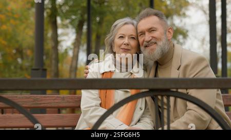 Caucasian happy couple mature bonding family sit on bench in city park old age love woman man relax enjoy weekend outdoors middle-aged male female Stock Photo
