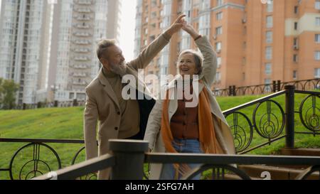 Carefree mature Caucasian couple grandparents family dancing romantic anniversary together dance in city park happy 60s aged older woman man moving Stock Photo