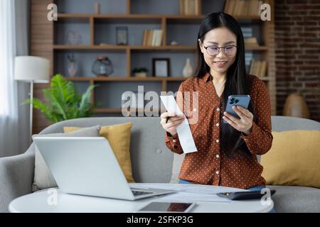 Asian woman managing personal finances from home with smartphone and laptop. Relaxed atmosphere conveys productivity, focus, and efficiency. Ideal for concepts of remote work and technology. Stock Photo