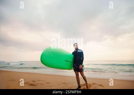 Young  bearded man with surfboard standing near a beach. Man with surfing board outdoors on a summer day. Stock Photo