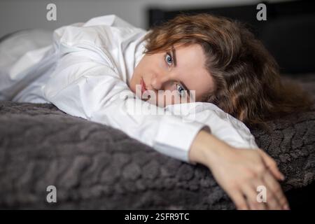 Serene young woman with blue eyes and long brown hair relaxes on a soft, gray blanket, wearing a crisp white shirt Stock Photo