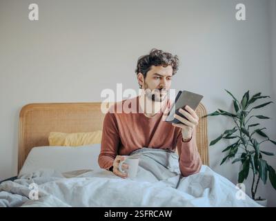 Young handsome middle eastern man reading on his tablet or ebook reader and enjoying a cup of coffee or tea. Morning ritual leisure relaxation. Stock Photo