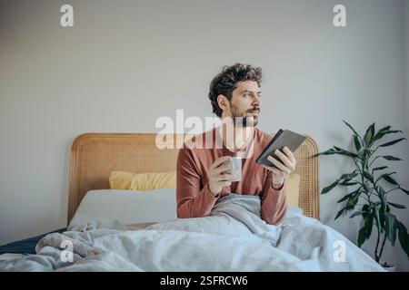 Young handsome middle eastern man reading on his tablet or ebook reader and enjoying a cup of coffee or tea. Morning ritual leisure relaxation. Stock Photo