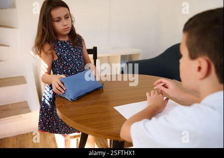 Two children are engaged in an activity involving school supplies at a wooden table, demonstrating curiosity and collaborative learning in a home or c Stock Photo