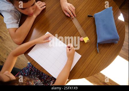 Two children collaborating on a written project at a wooden desk with tools like paper, a ruler, and a pencil case. Stock Photo