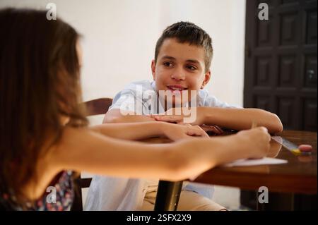 Two school-aged children interact during a study session at a table in a bright room. They seem engaged, highlighting elements of cooperation, educati Stock Photo