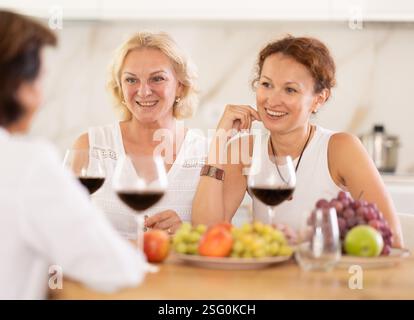 Relaxed adult female friends having fun at house party, sharing stories and laughing gathered around table with wine and appetizers in kitchen Stock Photo