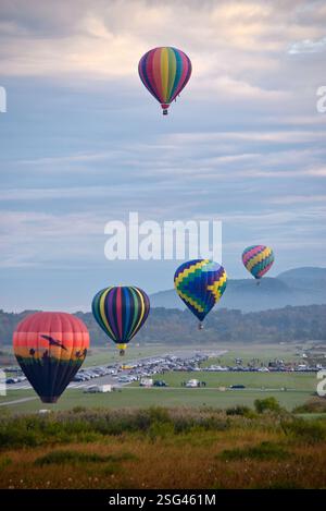 Colorful hot air balloons float above a lush landscape with a mountainous backdrop at the Adirondack Balloon Festival. Stock Photo