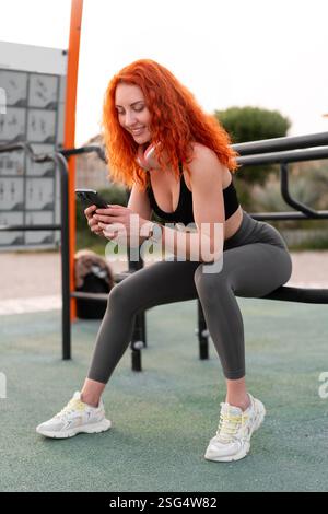 Smiling redheaded sporty woman in activewear using smartphone while sitting on equipment at outdoor gym. Happy female is relaxing after workout in park during daytime Stock Photo