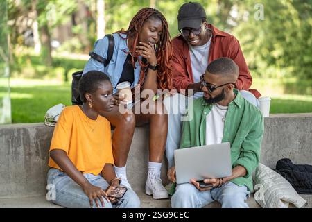 Cheerful group of African American friends drink coffee with laptop in campus park. Busy students Stock Photo