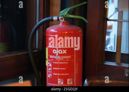A red Ajax fire extinguisher hangs next to a wooden door in Amsterdam, ready for any emergencies. Dutch text on the door adds a local touch. Fire safe Stock Photo