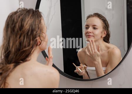 Morning beauty routine. Woman in a towel applying foundation in front of a mirror after a shower. Skincare, self-care, and natural makeup concept in a Stock Photo