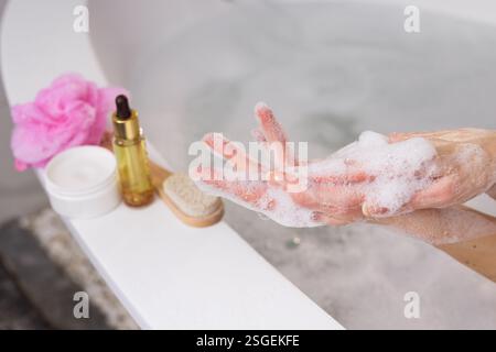 Relaxing spa moment. Woman washing hands with foamy soap near a bathtub. Skincare essentials like cream, oil, brush, and loofah on the side. Self-care Stock Photo