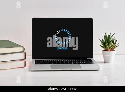 Modern laptop displaying loading screen on desk with books and plant, representing software update or data processing Stock Photo
