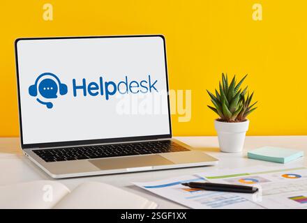 Modern laptop displaying helpdesk software interface on a desk with documents, charts, pen and plant, on a yellow background. Stock Photo