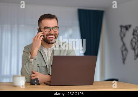 A smiling man talking on his phone and using a laptop while working at home. Stock Photo