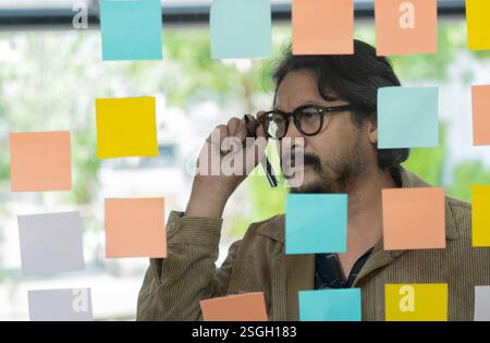 Serious thoughtful businessman ceo near glass board with adhesive sticky notes thinking about ideas of planning at office. Selective focus. Stock Photo