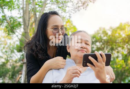 Smiling senior active with daughter sitting on the bench looking at smartphone. Using modern technology by elderly. Stock Photo