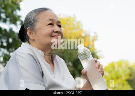 Smiling active senior woman holding water bottles, drink while relaxing in the park. Side view portrait. Stock Photo