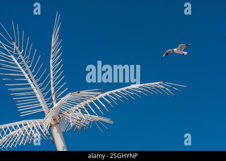 A low-angle shot of a metal sculpture resembling a palm tree, with a seagull flying in a clear blue sky. The image presents a contrast between artific Stock Photo