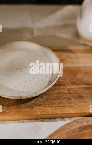 A white plate sits on a wooden cutting board. The plate is small and has a speckled design. The cutting board is made of wood and has a few scratches Stock Photo